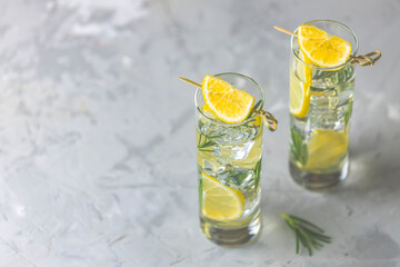 Two glasses of refreshing lemon lime drink with ice cubes in glass goblets against a light gray background. Summer fresh lemon soda cocktail with rosemary, selective focus