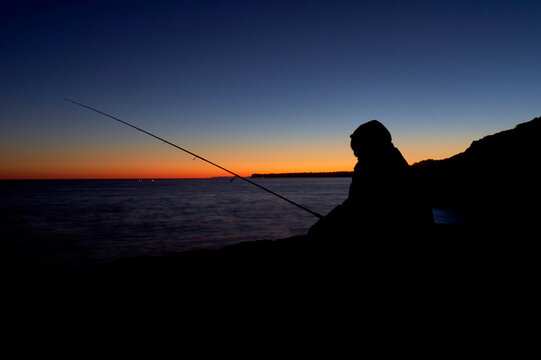 Silhouette of boy with fishing rod at calm sunset fishing off the cliff, long exposure photo