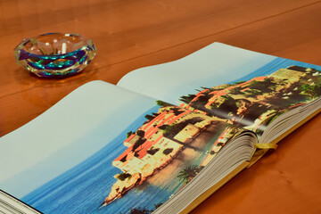 An open large book lies on a wooden table, and behind it is an ashtray with smoked cigars