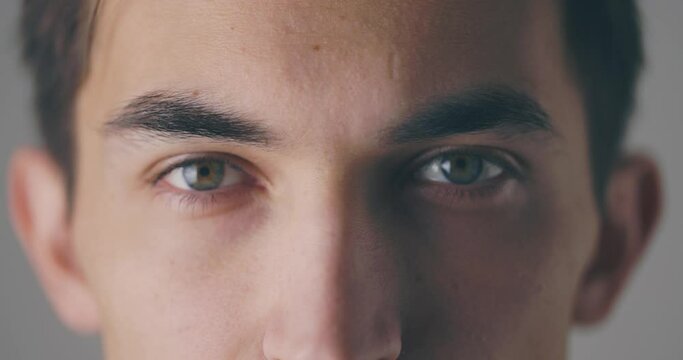 Close-up view of a calm young man is opening his eyes and looking to the camera at grey background wall in studio