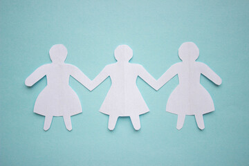 Figures of three girls in dresses holding hands, cut out of white paper. In the center of the photo on a blue background