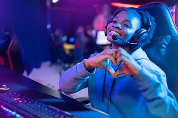 Streamer African beautiful girl shows heart sign with hands professional gamer playing online games computer, neon color