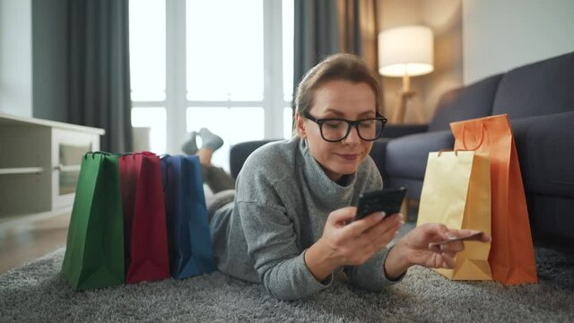 Happy woman with glasses is lying on the floor and makes an online purchase using a credit card and smartphone. Shopping bags around. Online shopping, lifestyle technology