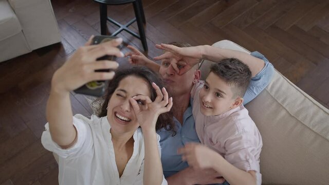 Young Mother Takes Pictures Of Her Family On The Phone. Mom Dad And Son On The Couch. They Show Funny Facial Expressions. Family Having Fun.