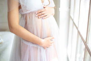 A pregnant woman in a white negligee touches her big belly with her hands. Light background. Waiting for a child. Side view. Close-up.