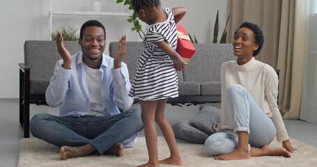 Three barefoot people family at home in living room interior, little generous Afro American girl with daughter holding red gift box behind back and holding out to dad makes surprise for Father's Day - Powered by Adobe