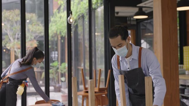Group of Asian restaurant staff in apron wearing face mask, cleaning and arranging the table with social distancing concept, preparing for reopen after lockdown due to coronavirus pandemic. 