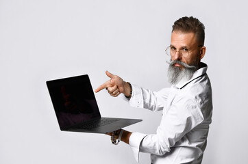 Stylish grey-haired bearded hipster man in eyewear pointing to black blank laptop screen looking at camera. Studio shot closeup portrait isolated on white background. Recommend, presentation
