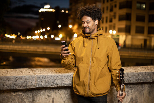 Man with guitar using mobile phone while standing by retaining wall