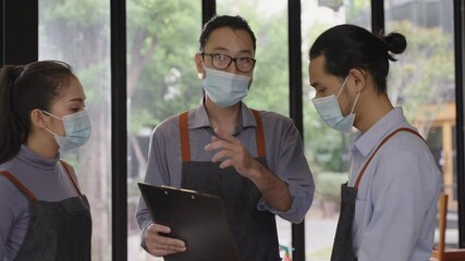 Asian male restaurant manager wearing face mask, explaining and assigning jobs to his colleagues, preparing for reopen after lockdown due to coronavirus pandemic with new normal business concept.
