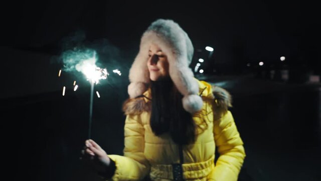 Young cute girl in a winter fur hat and yellow jacket in a festive mood. Sparkling burning sparklers in hand with flying sparks. Dancing and having fun. Holidays concept. Moving camera