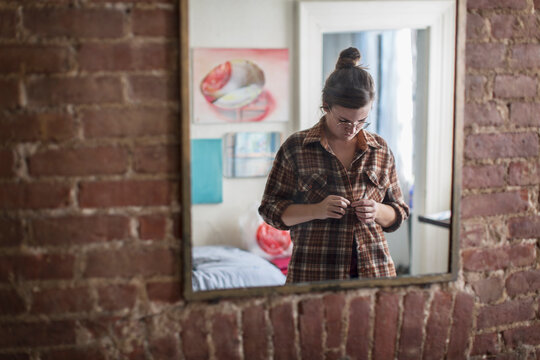 Young woman buttoning shirt in bedroom mirror