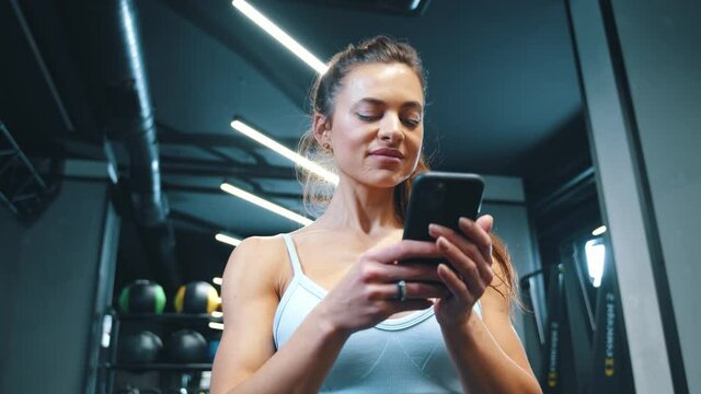 Fitness attractive woman using smartphone social media network notification user interface application in modern professional gym indoors.