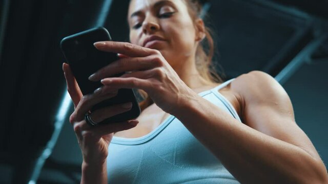 Young attractive fitness woman in gym overalls using mobile phone social network typing messages on smartphone screen standing in sports club.