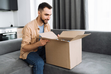 Home delivery. Stylish caucasian guy got his order, unpack a parcel. Happy man with interest look inside cardboard box, sitting on sofa, the guy shopping in internet stores, buying new clothes online