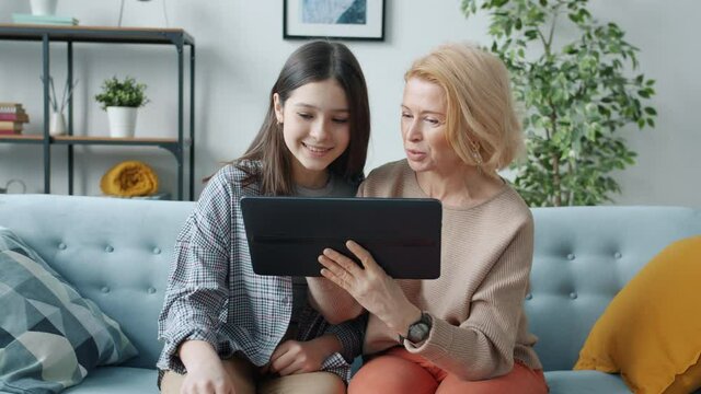 Adult mother and teenage daughter are using tablet touching screen and talking sitting on couch at home. Parenthood and family relations concept.