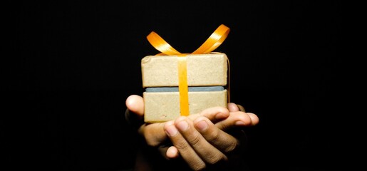 Closeup of Human hand holding / Giving / receiving Christmas Gift boxes wrapped with orange ribbon in a black isolated background with copy space for texting