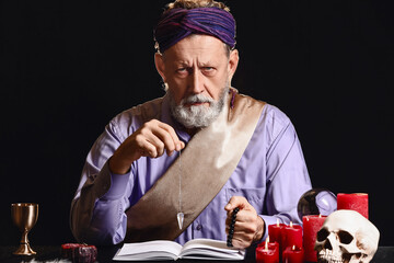 Male fortune teller with pendulum and book at table on dark background