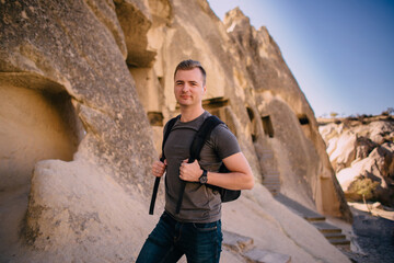 A young male tourist with a backpack stands on a background of rocks and looks at the camera.