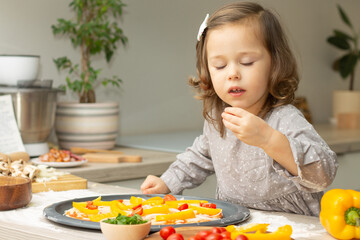 Cute little girl 2-4 in gray dress cooking pizza in kitchen. Kid arranges ingredients on pizza base