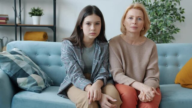 Portrait of mature woman and teenage girl looking at camera with sad faces expressing family crisis sitting on couch together. Relations and emotions concept.