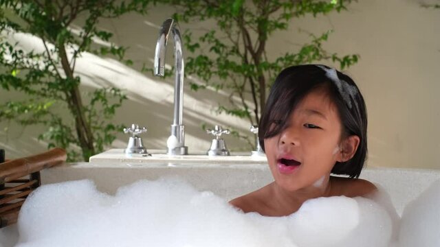 Happy asian girl take bublble bath and play with soapy foam and blowing foam at bathtub. Kid having fun with foam in bath.