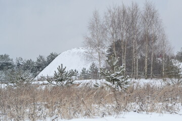 Field in winter with snowy hill against sky backgdrop. Winter rural landscape with big white hill, snow field and gray cloudy sky