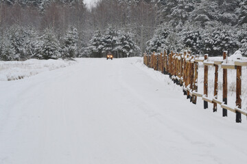 Winter Road In Snowy Forest near snowy wooden fence, Far away light Car headlights