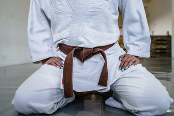 Close up on midsection of unknown woman in white brazilian jiu jitsu or judo kimono gi sitting on tatami mats wearing brown belt on training - Powered by Adobe