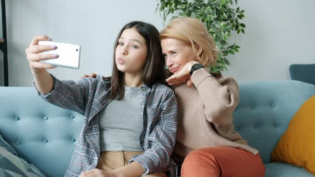Teenage girl taking selfie with mother having fun making funny faces using smartphone camera indoors in apartment. Family relations and technology concept.