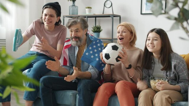 Parents and children are enjoying football game on TV cheering doing high-five holding US flag and ball sitting on couch at home. People and sports concept.