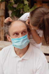 Stock Photo - Little granddaughter in a face mask hugs and wants to protect grandfather from an epidemic.  Family support during quarantine isolation due to outbreaks of coronavirus. Elderly at risk