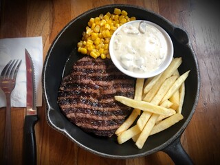 Grilled sliced of well done beef steak with mushroom sauce, French fries and sauce corn on hotplate pan. Fork and spoon side the table. Flat lay style