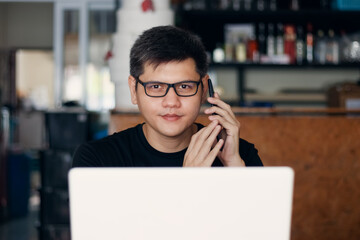 A young businessman talking on the phone with clients about business planning and financial and investment planning, using a laptop to work in a coffee shop.