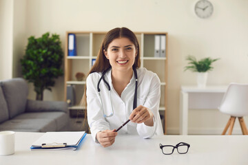 Smiling female doctor looking at camera sits by table. Doctor's office and hospital interior.