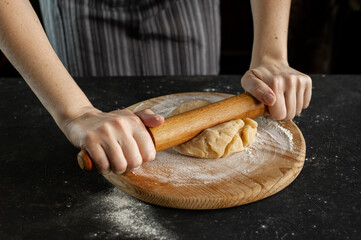Recipe of cookies. Women's hands roll out the dough with a rolling pin on a wooden board on a dark table.