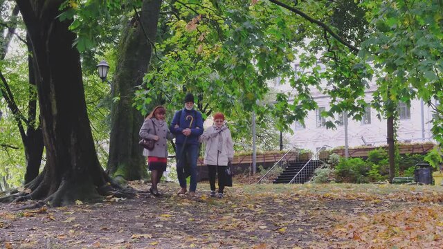 Stylish retired man holding umbrella walks arm in arm with elegant women friends wearing warm coats along cold autumn park