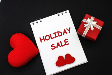 On a black background among a red heart and a gift box there is a notebook with the inscription holiday sale. The concept of holiday sales