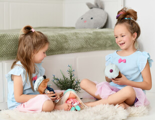Happy laughing kid girls sisters in home dresses spend time together talking and playing funny games with their dolls. Happy childhood, cheerful lifestyle, games, comfortable pastime, hobby concept