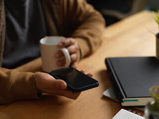 Male hand holding smartphone and coffee cup while sitting at workplace