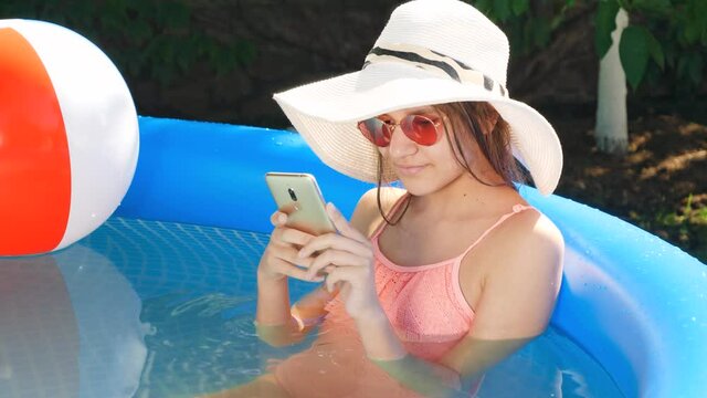 Portrait of beautiful young girl relaxing in outdoor swimming pool at backyard and typing message on smartphone. Concept of happy summer holidays and vacation