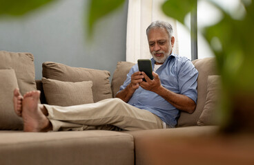 A SENIOR ADULT MAN SITTING COMFORTABLY ON SOFA AND USING MOBILE PHONE	