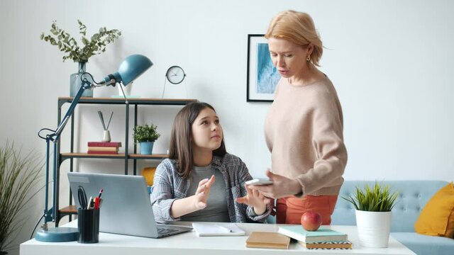Teen girl playing video game instead of doing homework then fighting with mother while mature woman asking her to stufy. Teenage lifestyle and gadgets concept.