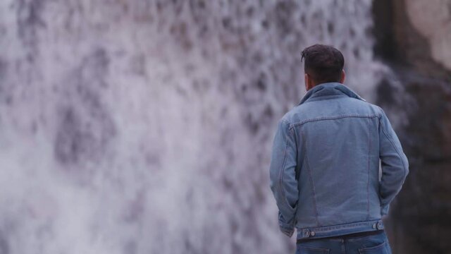 Young Indian Man with hands in pocket looking with confidence at waterfall, wearing blue denim jacket and blue jeans. Mid Shot, blurred background, looking left to right