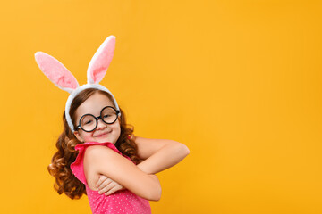 Funny happy cute little girl wearing Easter bunny ears and glasses on a yellow background. The child hugs himself with his arms.