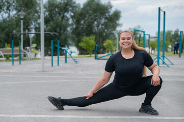 Obese young caucasian woman doing side lunges. Fat beautiful smiling girl in a black tracksuit is engaged in fitness outdoors. Warming up before training.