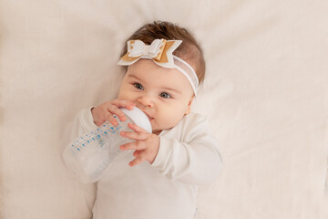 baby six months lies in a crib with a bottle and drinks water in a white bodysuit
