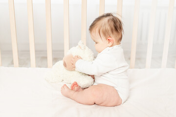 baby six months playing in a crib in a white bodysuit with a Teddy bear