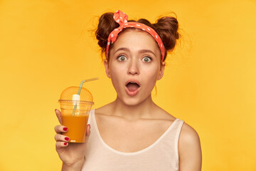 Portrait of attractive, red hair girl with red doted hairband. Wearing white shirt and holding her smoothie, expressing shock. Watching at the camera isolated over yellow background