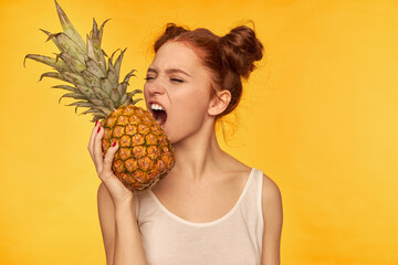 Young lady, pretty ginger woman with two buns. Wearing white shirt and trying to bite pineapple...